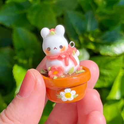 A small decorative figurine of a white bunny sitting on an orange pot with a floral pattern, set against a leafy background.